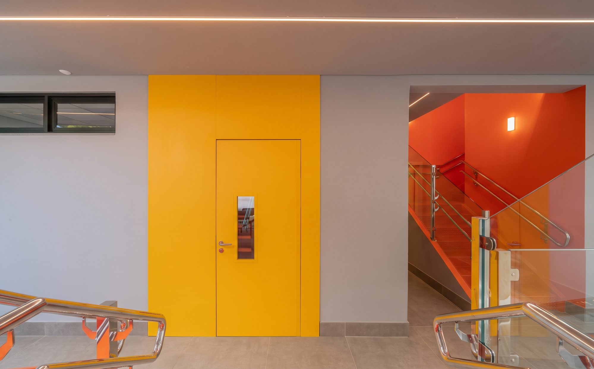 Porta amarela em revestimento PET numa escola, ao lado da escada laranja.